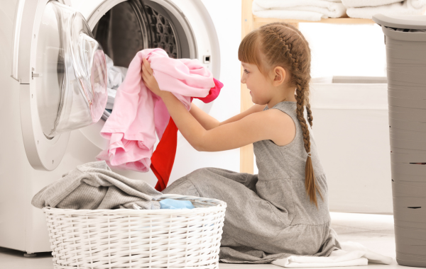 A little girl is seated in front of a dryer removing the clean clothes and putting them in a basket