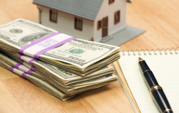 A stack of hundred dollar bills is pictured in front of a model of a house. There is a pad of paper and a pen to the right of the money