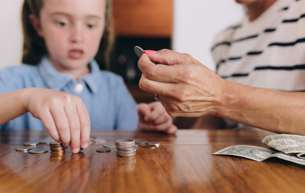 A child is counting coins, and in the foreground of the image, an adult's hand is holding out a quarter