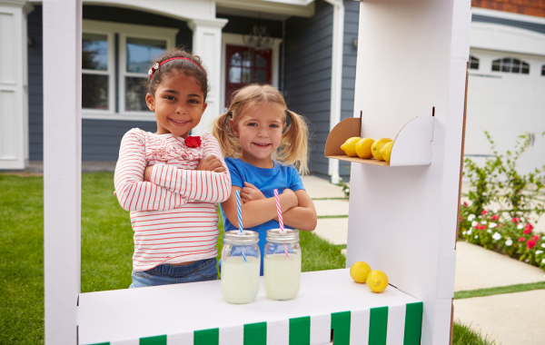 Picture of two young girls standing proudly behind their lemonade stand. There are two glasses of lemonade in front of them.
